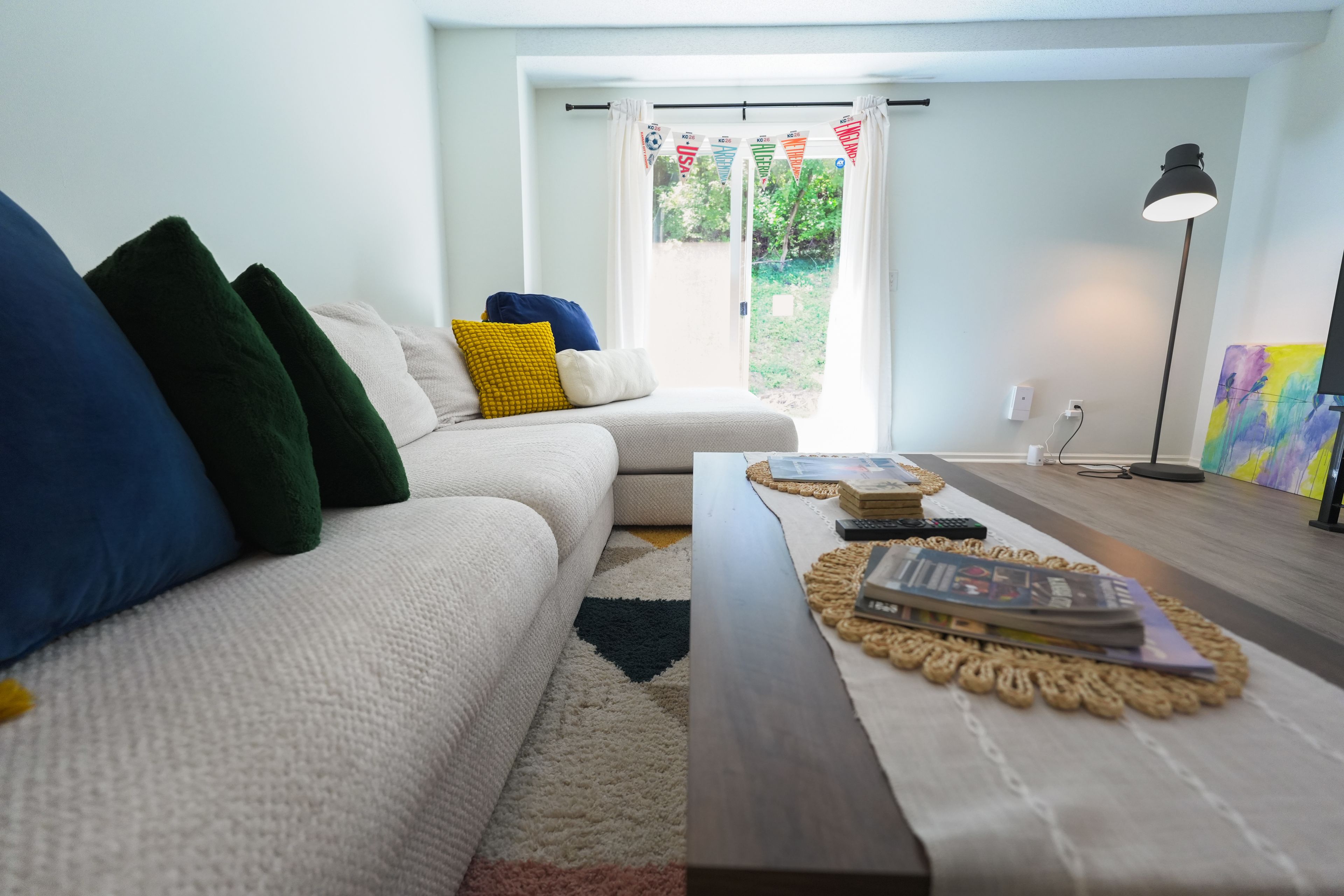 Ein Wohnzimmer mit einem weißen Sofa, bunten Kissen, einem Couchtisch mit Büchern, einer Lampe und Blick durch ein Fenster.
