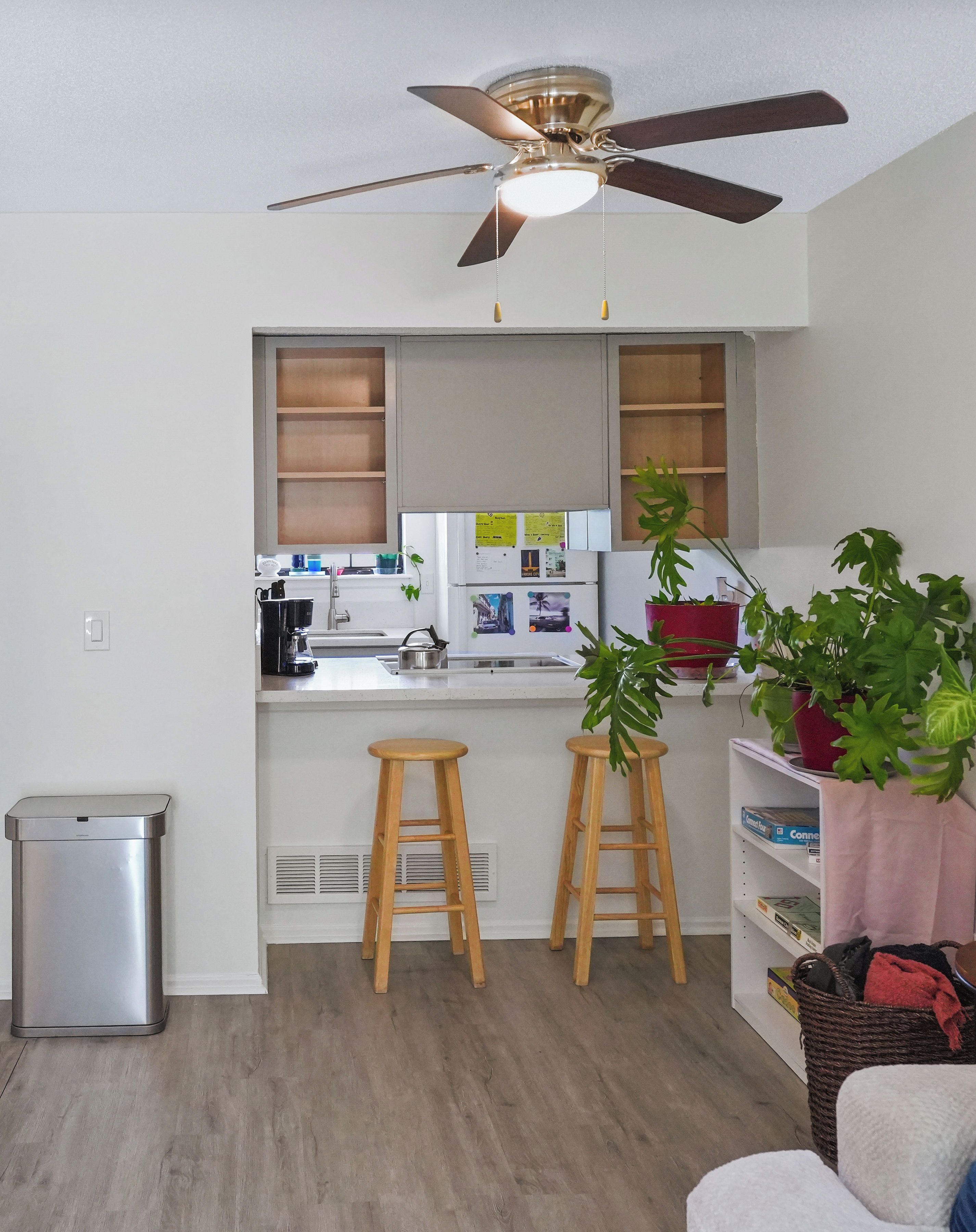 A compact kitchen with a ceiling fan, a small trash bin, wooden stools, and potted plants.