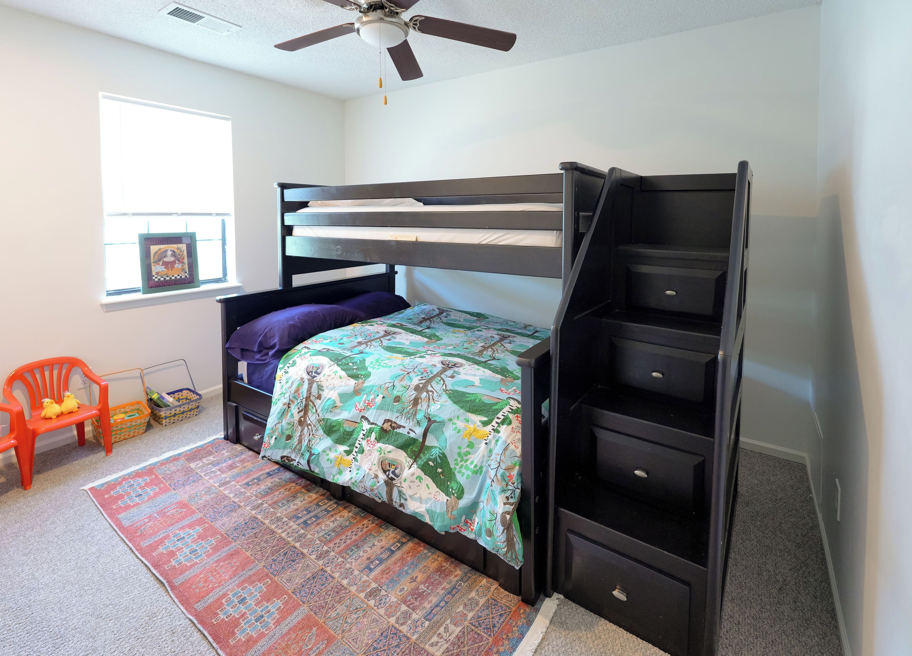 A bunk bed with a built-in desk and shelving, a window with blinds, a colorful rug, and a ceiling fan.