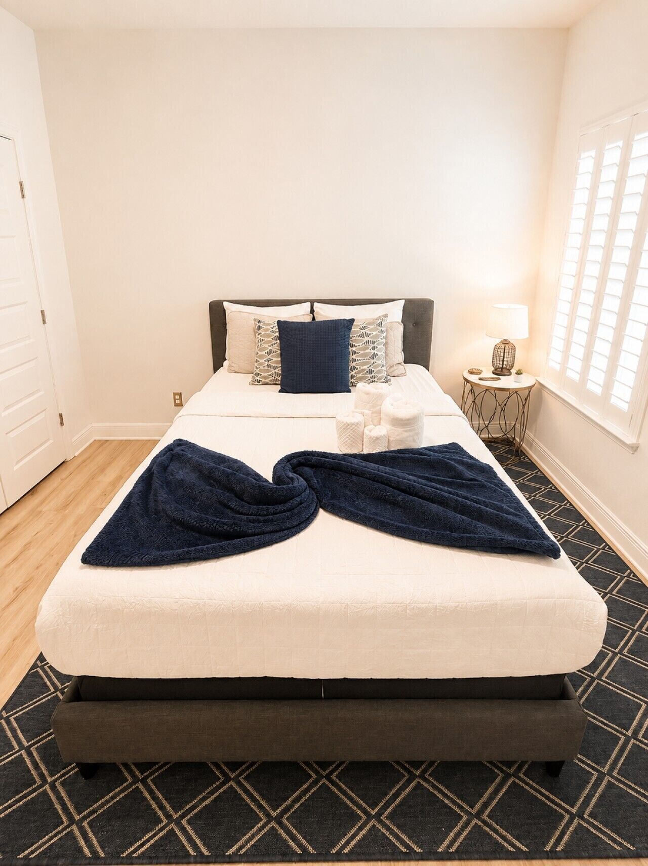 A neatly made bed with a dark blue blanket, a bedside table with a lamp, and a window with blinds.