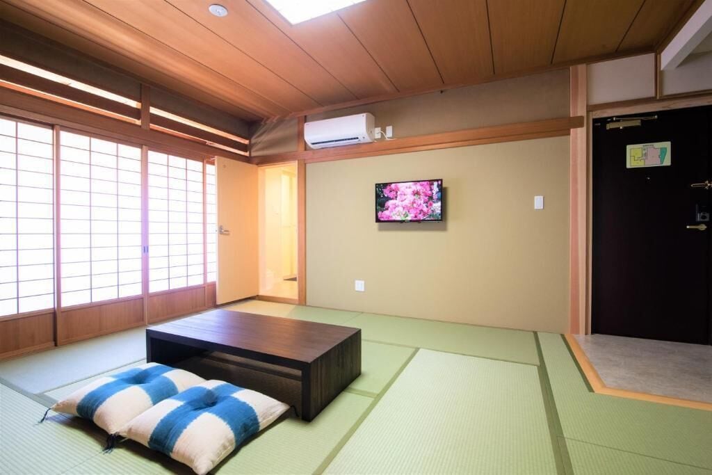 A traditional Japanese room with tatami flooring, a tatami mat bed, a television on the wall, and a sliding door.