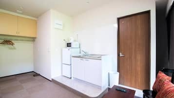 A compact kitchen with a microwave, sink, and cabinets, a wooden door, and a small table with a remote control.