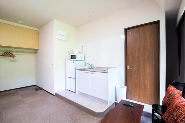 A compact kitchen with a microwave, sink, and cabinets, a wooden door, and a small table with a remote control. - Arbor house (Kagoshima , Nishida)