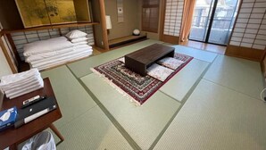 Une pièce avec un sol recouvert de tatamis, un futon servant de lit, une petite table avec une télécommande et une porte coulissante donnant sur un balcon.