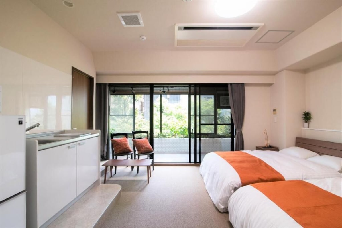 Een hotelkamer met kitchenette, een bed met oranje accentkleur, een zithoek met stoelen en een groot raam met uitzicht.