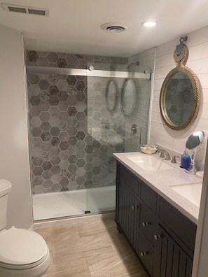 Et badeværelse med et brusebad i glas, et hvidt toilet, et badeværelsesbord med to vaske og et spejl.