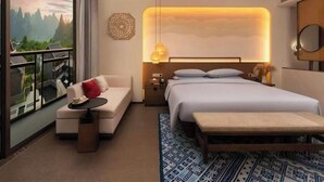 Quarto de hotel com cama, sofá, mesinha, mesa de cabeceira, espelho e vista para uma paisagem montanhosa através de grandes janelas.