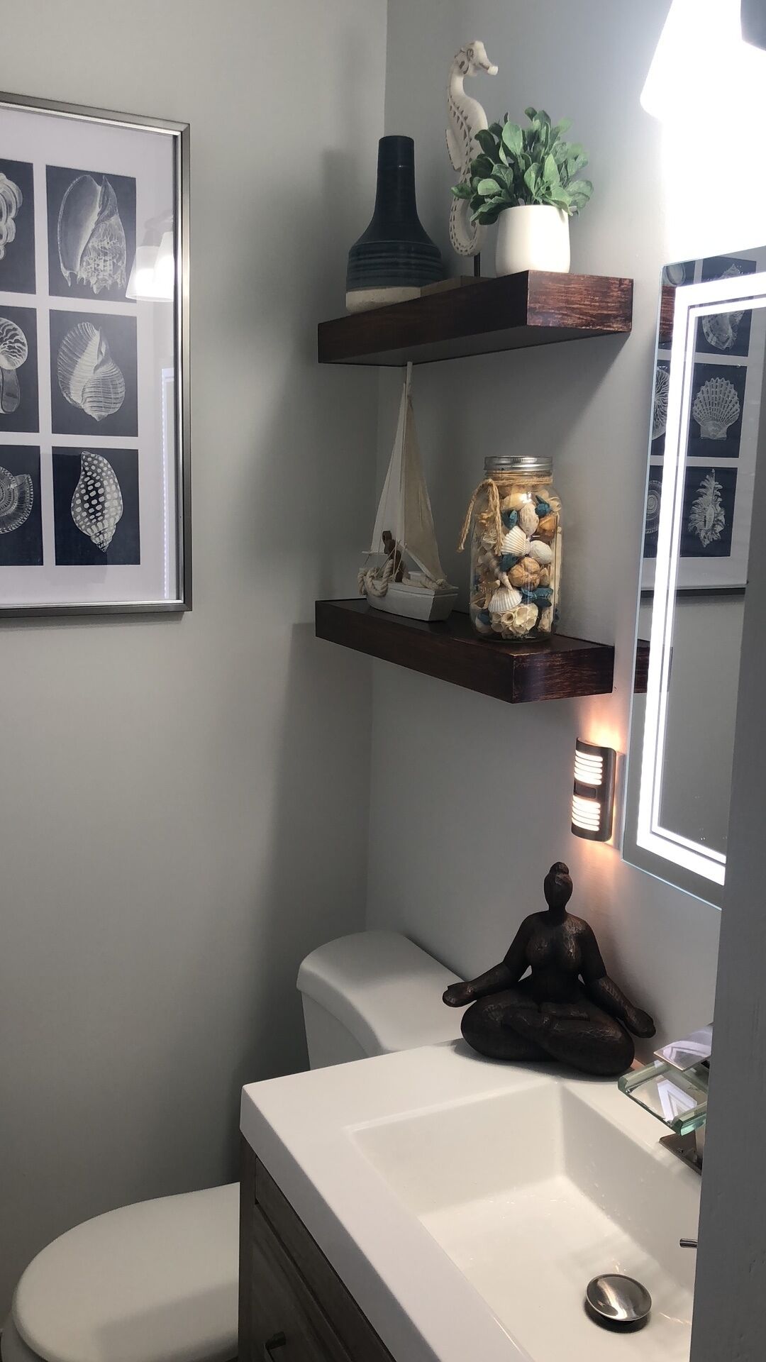 A bathroom with a white sink, a toilet, a framed seashore print, a decorative seahorse, a potted plant, a jar of seashells, and a small statue.
