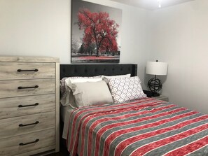 A bedroom with a bed, a nightstand, a lamp, a dresser, and a wall-mounted picture.