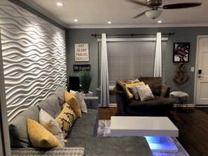 A modern living room with a grey sofa, a brown armchair, and a white coffee table. There are decorative pillows and a plant on the sofa.