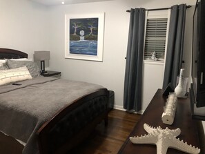 Een slaapkamer met een bed, een nachtkastje, een lamp, een lijst met een schilderij, een raam met gordijnen, en een decoratie van een zeester.