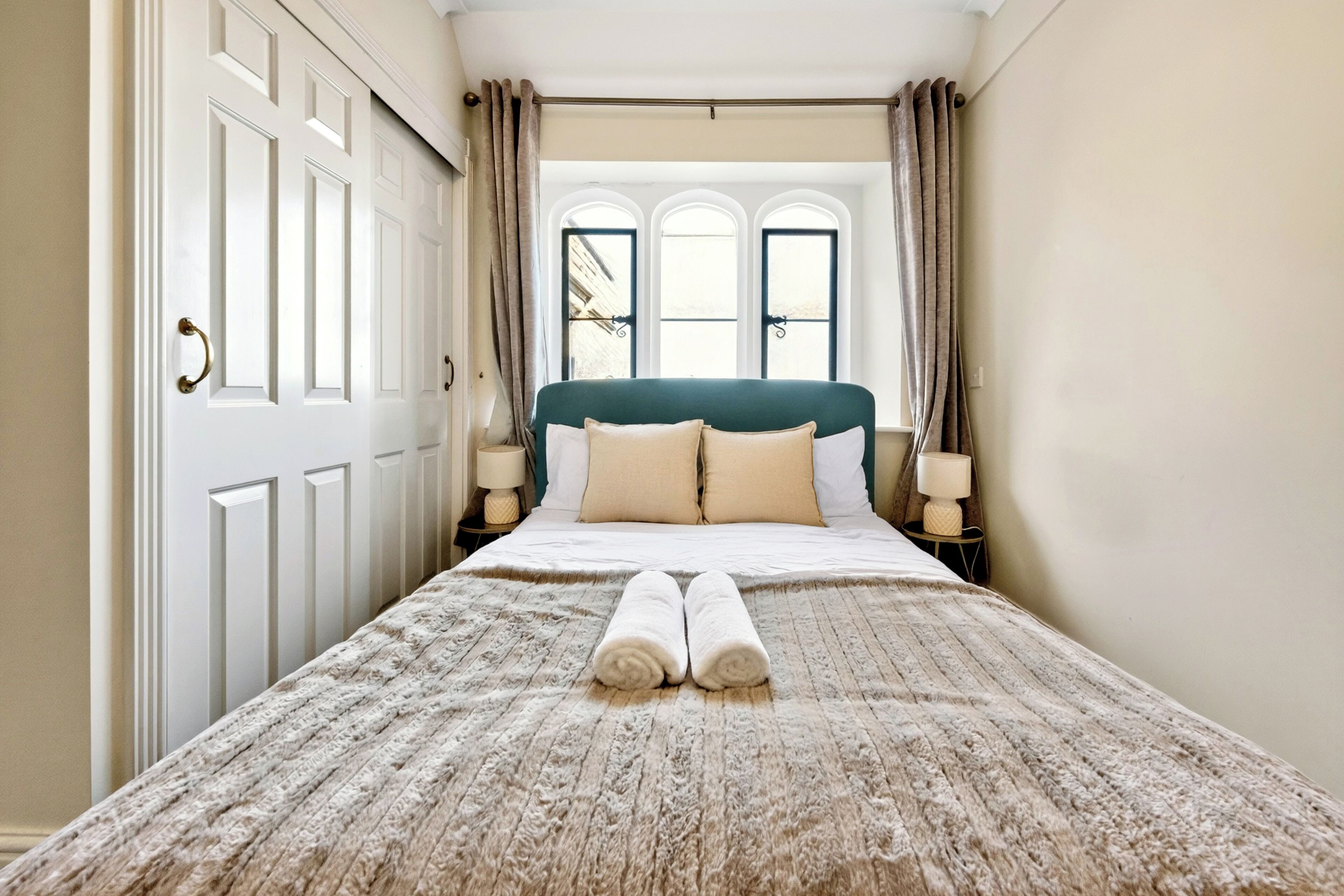 A hotel room with a large bed, two bedside lamps, a window with curtains, and a door with a handle.