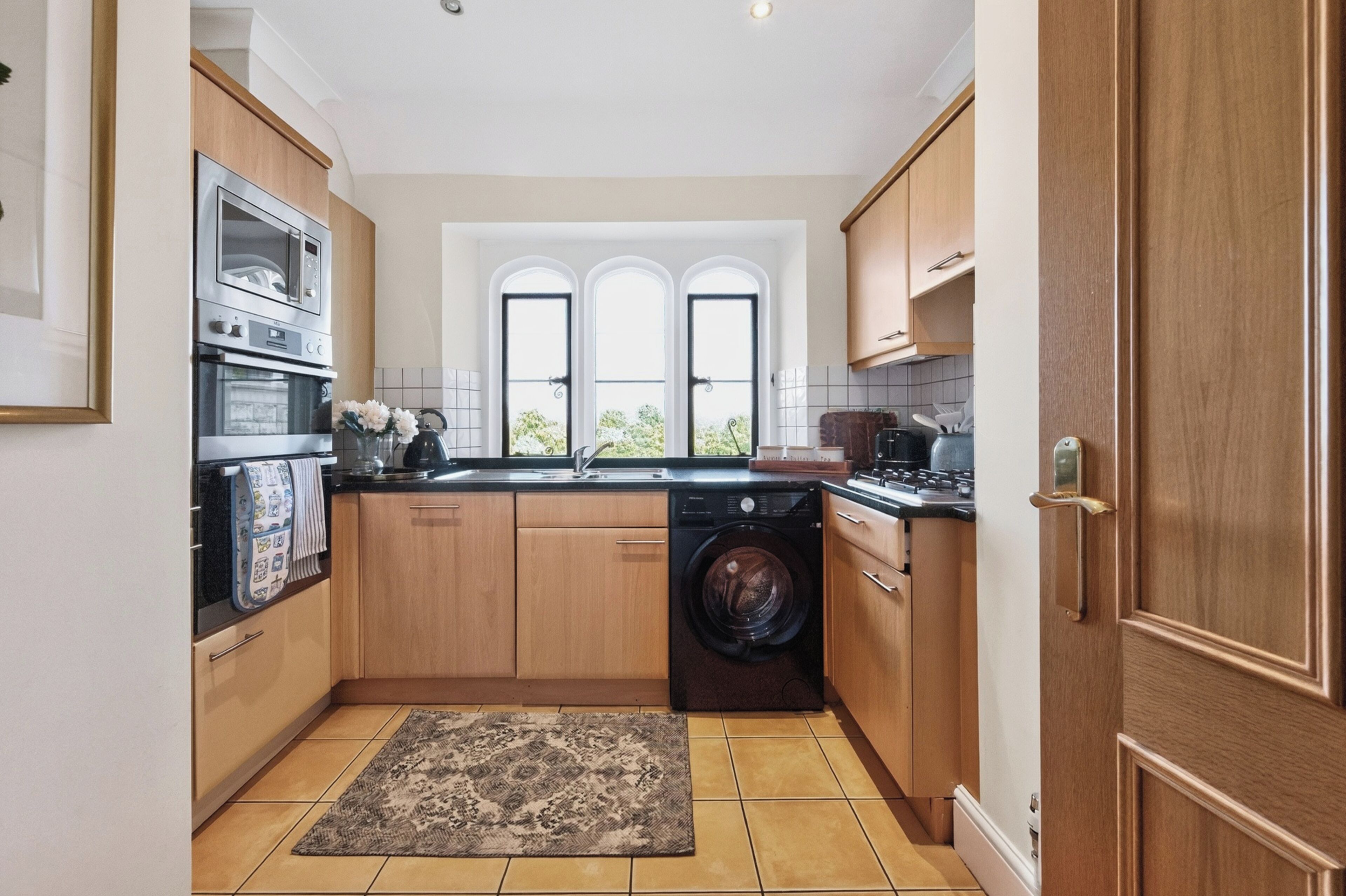 A compact kitchen with wooden cabinets, a built-in oven, a washing machine, and a window with a view.