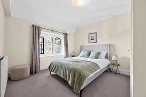 Un dormitorio con una cama, una ventana con cortinas, una mesita de noche con una lámpara y un cuadro en la pared.