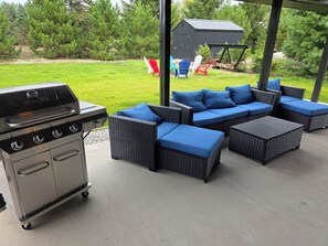 Un patio con una parrilla, muebles de exterior y vistas a un césped y árboles.