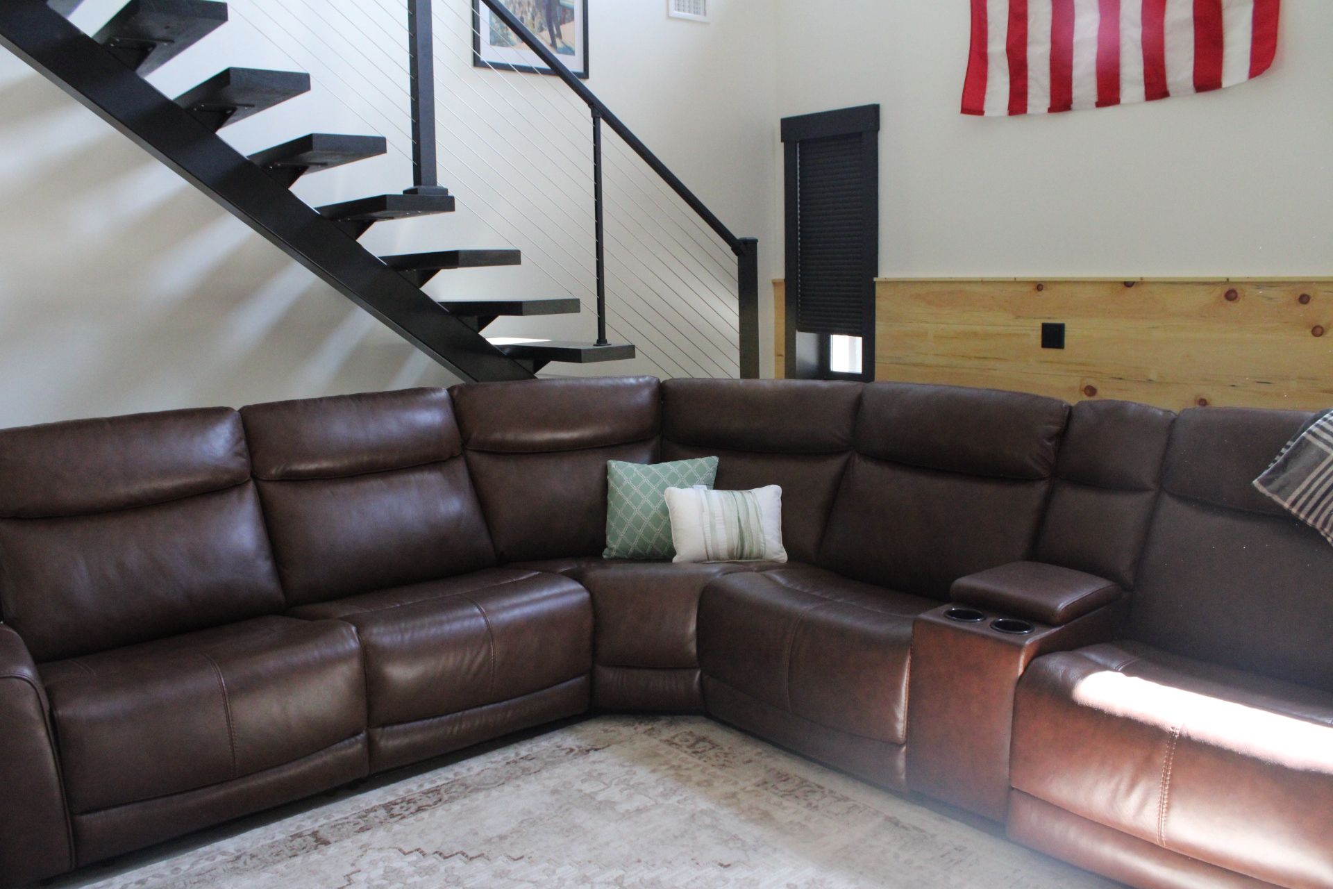 A corner living room with a brown leather sectional sofa, a wooden floor, a staircase, and a red and white striped flag on the wall.