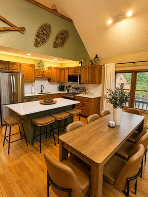 A kitchen with wooden cabinets, a stainless steel refrigerator, a large island with stools, and a dining table with chairs. - Schuss Haus • Modern Chalet • Hot Tub • Ski • Golf • A/C (Bellaire)