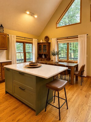 A kitchen with a large island, wooden cabinets, and a dining area with a view of trees. - Schuss Haus • Modern Chalet • Hot Tub • Ski • Golf • A/C (Bellaire)