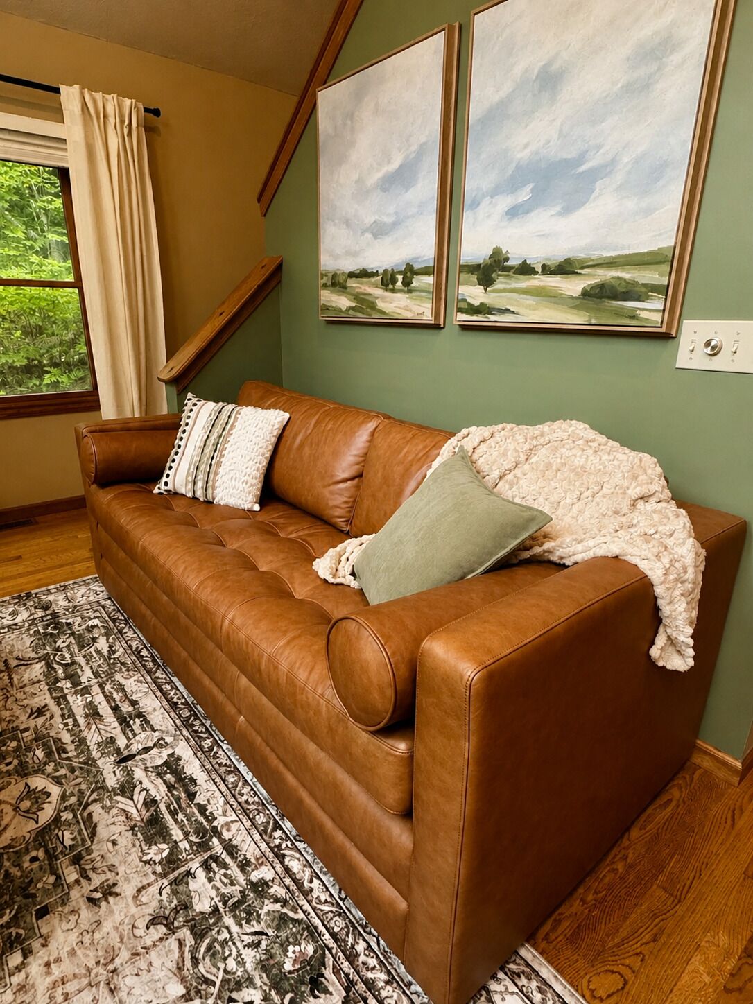 A brown leather sofa with cushions, a patterned rug, and two landscape paintings on the wall.