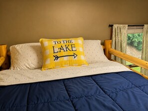 Un letto con una coperta azzurra, due cuscini con motivi ad ancora e un cuscino giallo con la scritta 'TO THE LAKE'.