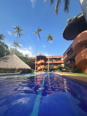 A swimming pool with a clear blue water surface, surrounded by palm trees and a building with a red facade. - 3-bedroom condo 1 Block from Playa La Ropa (Ixtapa Zihuatanejo)