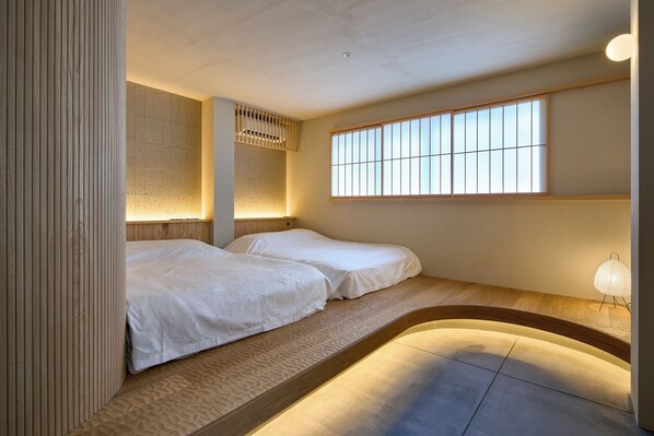 A room with two beds, a window with vertical blinds, and a wall-mounted light fixture.