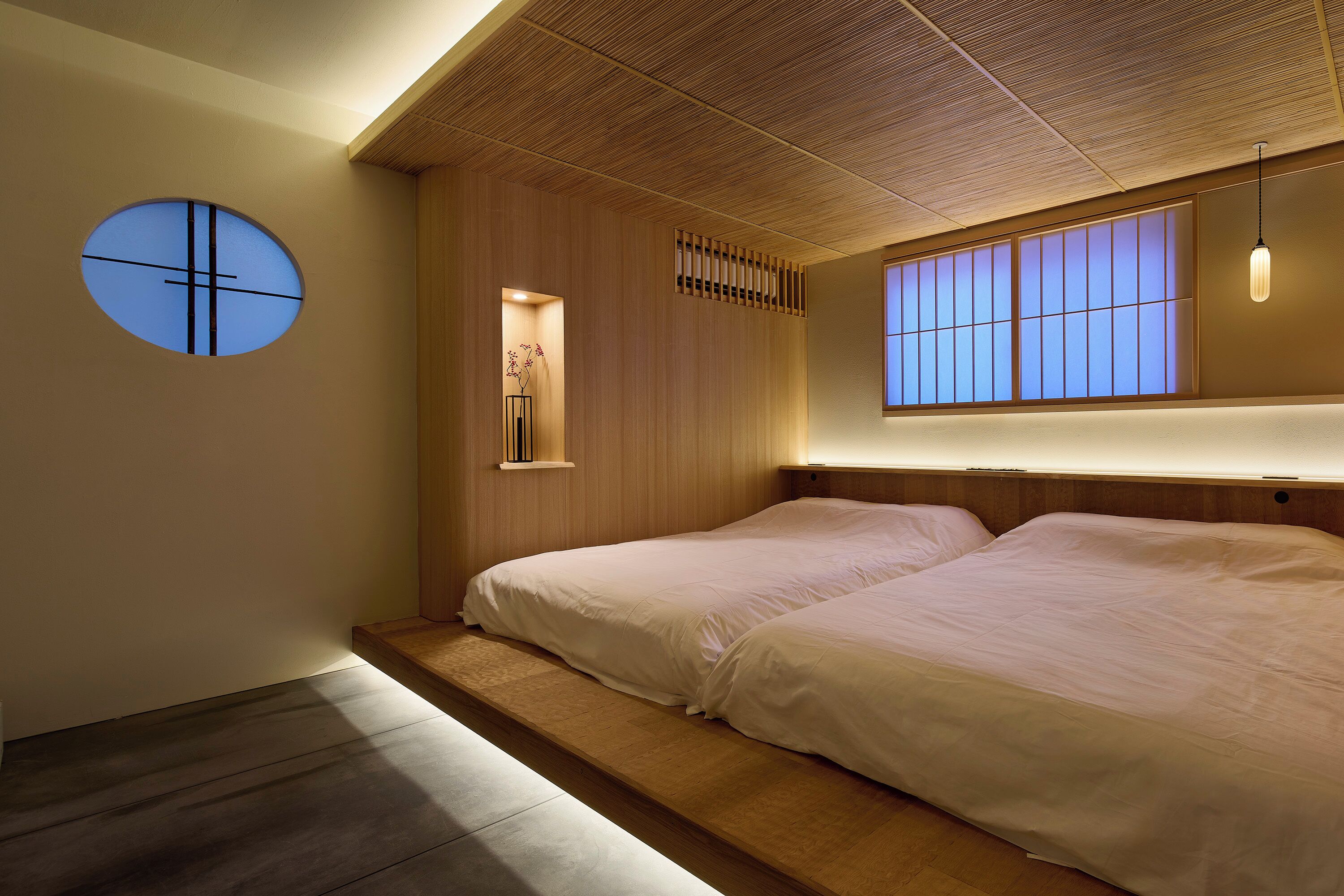 A hotel room with two beds, a wooden ceiling, a small window, and a wall-mounted shelf with a plant.