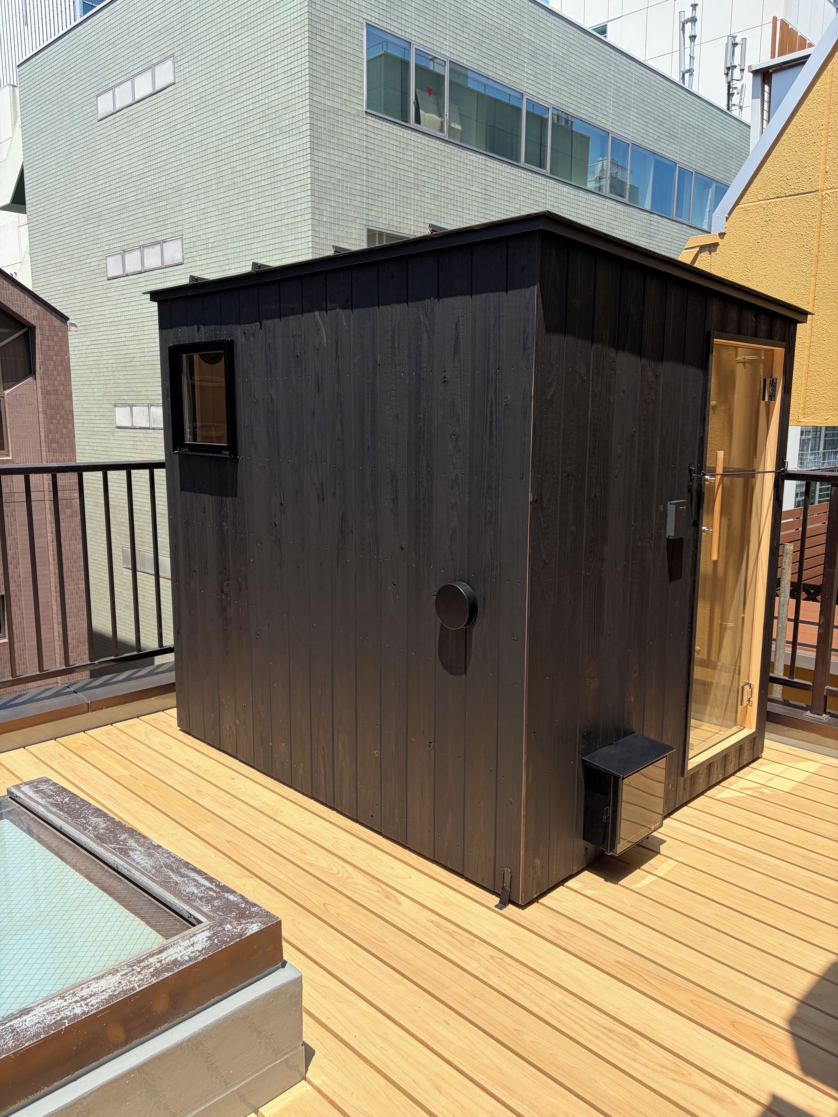 A wooden deck with a sauna, a wooden structure with a door, and a building in the background.