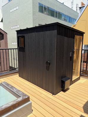 A wooden deck with a sauna, a wooden structure with a door, and a building in the background.