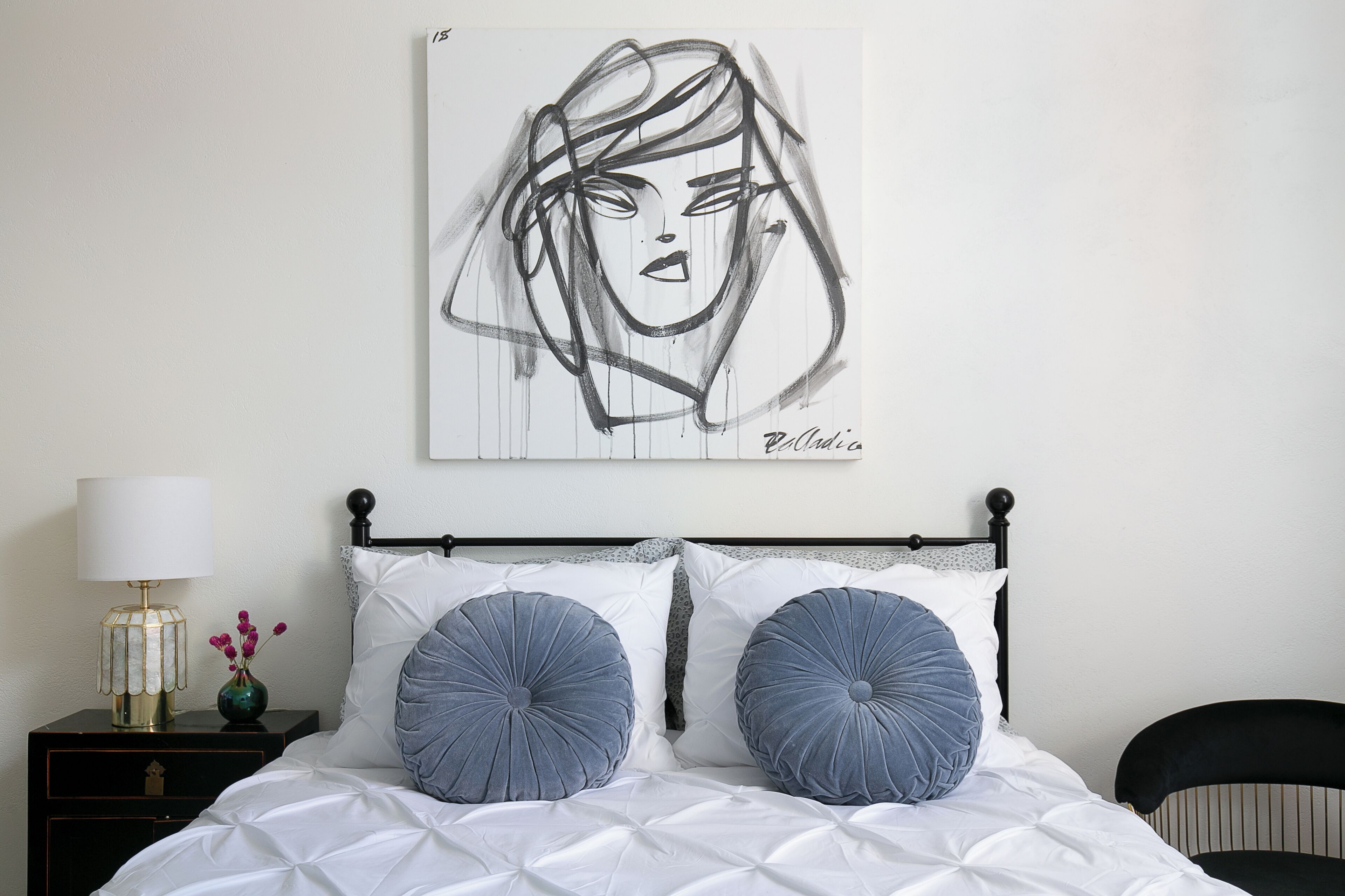 Une chambre à coucher comprenant un lit, deux coussins décoratifs, une table de chevet avec une lampe et une œuvre d’art accrochée au mur.