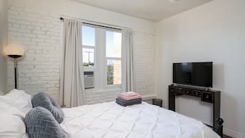 Ett hotellrum med en vit tegelvägg, en säng med vita sängkläder, en platt-TV som är monterad på väggen och ett fönster med gardiner.