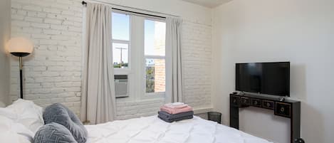 A hotel room with a white brick wall, a bed with white bedding, a flat-screen TV mounted on the wall, and a window with curtains.