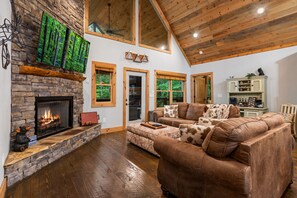 A spacious living room with a stone fireplace, a large window, and a cozy seating area. - Modern Cabin - New Hot Tub, Fire Pit, Arcades, Games, Wi-Fi - Mins to Downtown! (McCaysville)