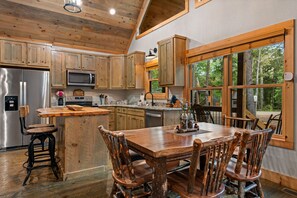 A rustic kitchen with wooden cabinets, a stainless steel refrigerator, a wooden dining table with chairs, and a large window with a view of trees. - Modern Cabin - New Hot Tub, Fire Pit, Arcades, Games, Wi-Fi - Mins to Downtown! (McCaysville)