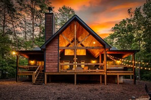 A cabin with a wooden exterior, a chimney, a deck with seating, and string lights. - Modern Cabin - New Hot Tub, Fire Pit, Arcades, Games, Wi-Fi - Mins to Downtown! (McCaysville)