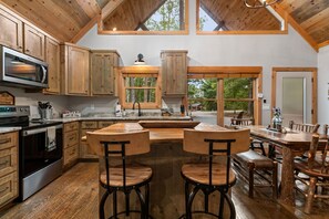 A spacious kitchen with wooden cabinets, a large island with bar stools, and a view of the outdoors through large windows. - Modern Cabin - New Hot Tub, Fire Pit, Arcades, Games, Wi-Fi - Mins to Downtown! (McCaysville)