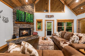 A cozy living room with a stone fireplace, a large window, and a wooden ceiling. - Modern Cabin - New Hot Tub, Fire Pit, Arcades, Games, Wi-Fi - Mins to Downtown! (McCaysville)
