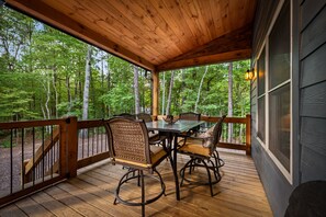 A wooden deck with a table and chairs, surrounded by a railing, overlooking a forested area. - Modern Cabin - New Hot Tub, Fire Pit, Arcades, Games, Wi-Fi - Mins to Downtown! (McCaysville)