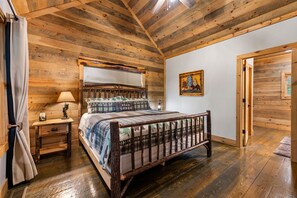 A wooden-paneled bedroom with a bed, a nightstand, a lamp, and a door. - Modern Cabin - New Hot Tub, Fire Pit, Arcades, Games, Wi-Fi - Mins to Downtown! (McCaysville)
