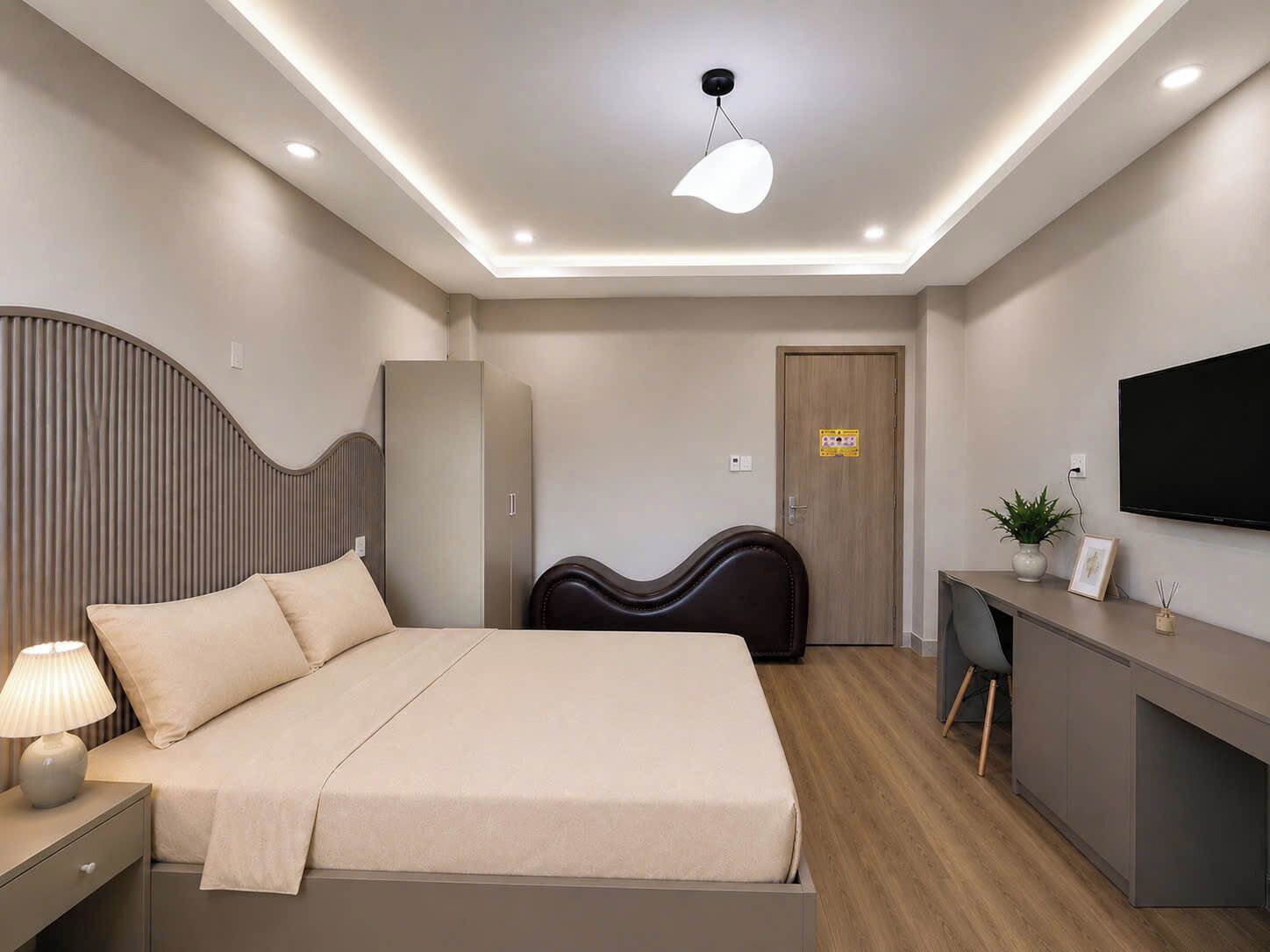 Habitación de hotel moderna con cama, mesita de noche, lámpara, armario, escritorio, silla, televisor y una planta en maceta.
