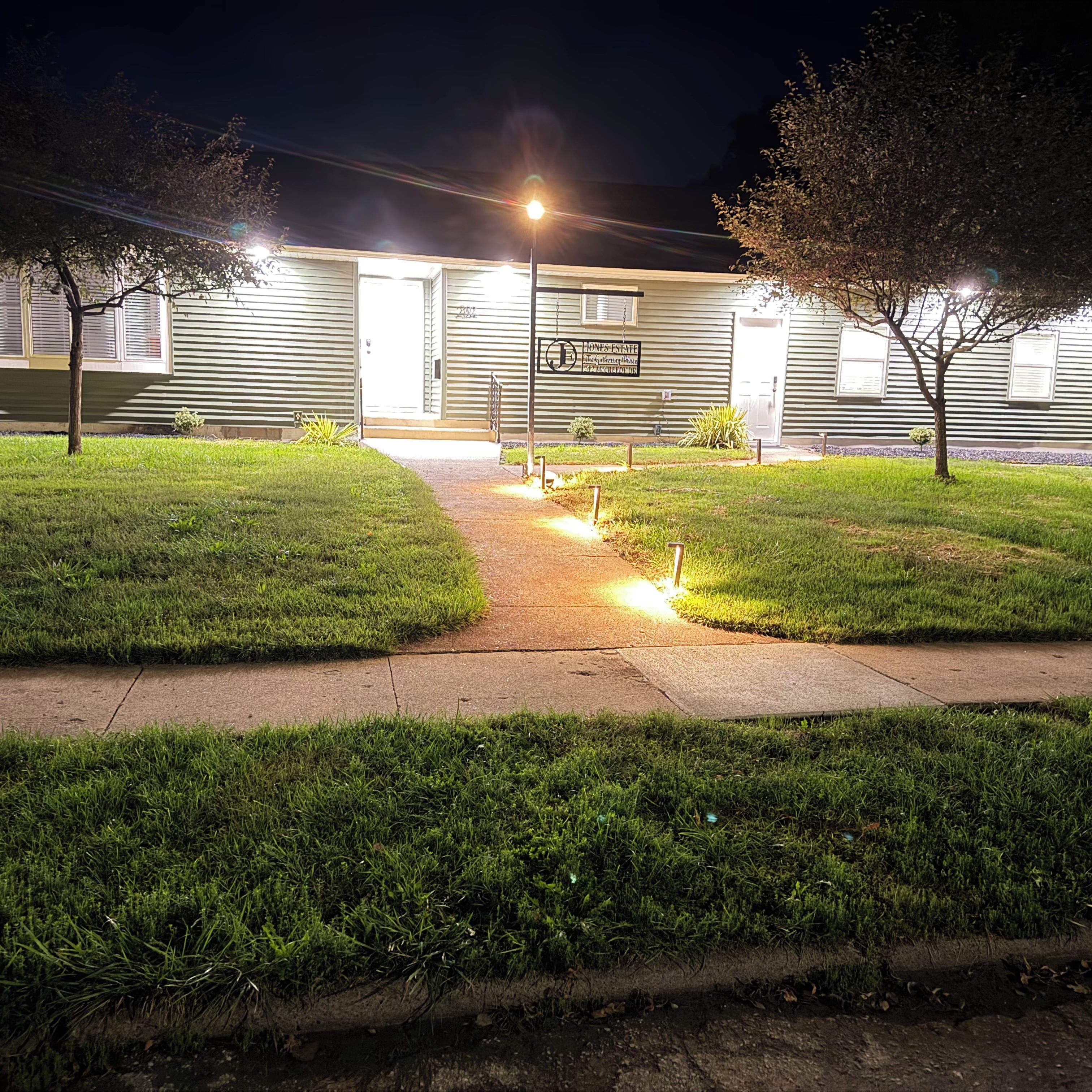 A gated residential property at night with a well-lit entrance, a paved driveway, and a grassy lawn.