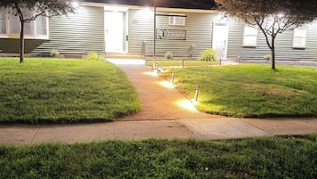 A gated residential property at night with a well-lit entrance, a paved driveway, and a grassy lawn.