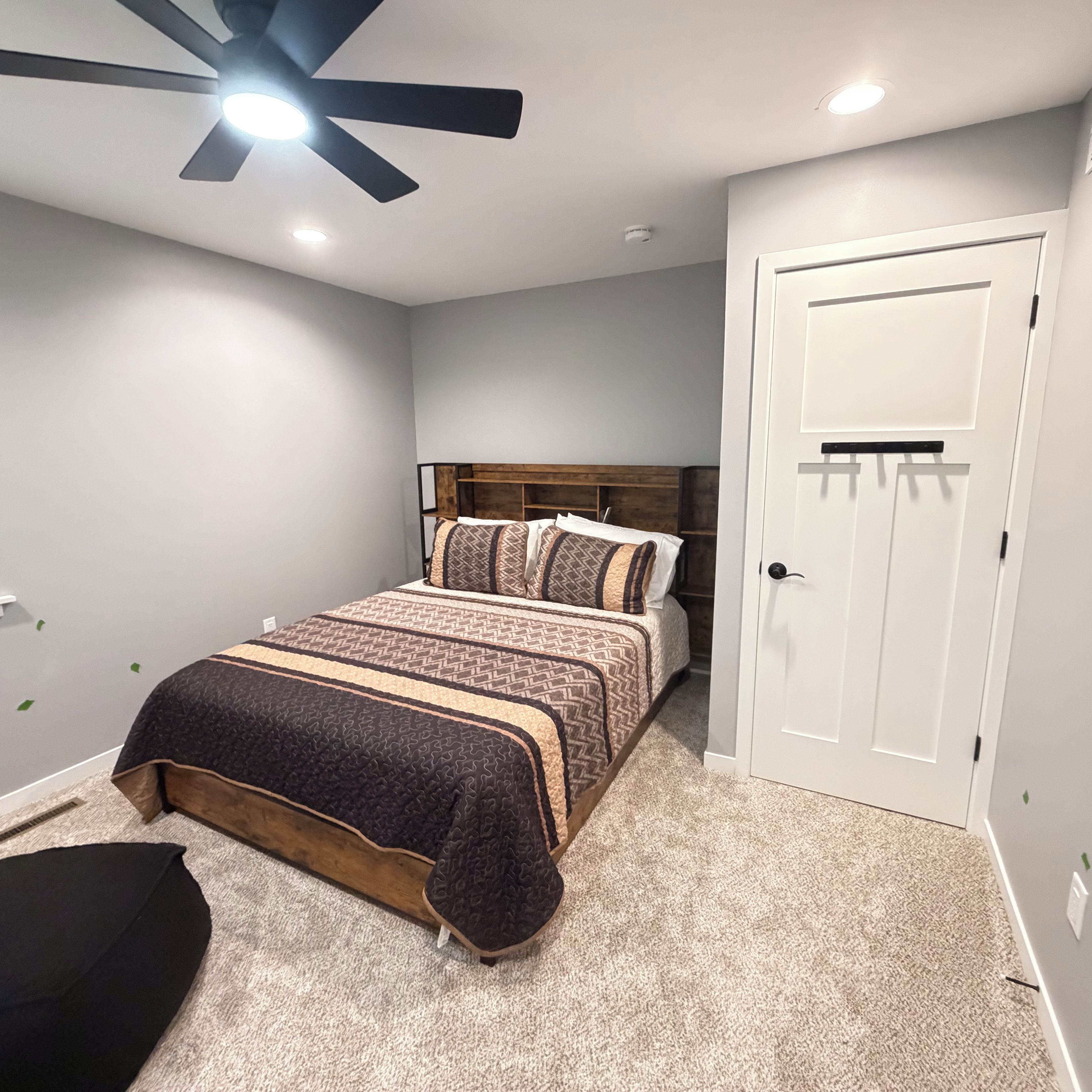 A bedroom with a bed, a ceiling fan, a door, and a carpeted floor.