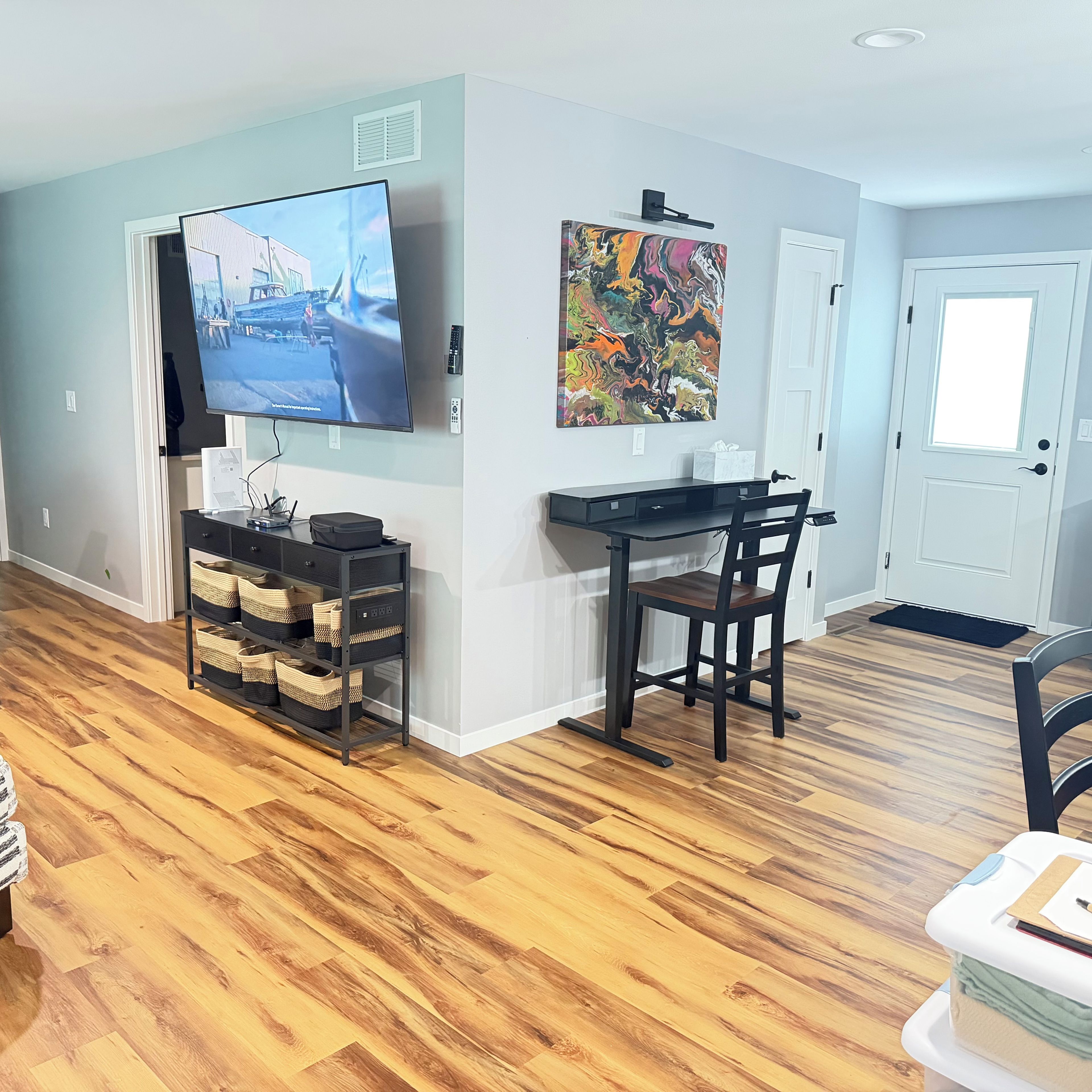 A living room with a flat-screen TV, a painting, a desk with a chair, and a wooden floor.