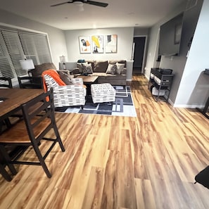 A living room with a sofa, coffee table, and ottoman, a dining table with chairs, and a TV mounted on the wall.