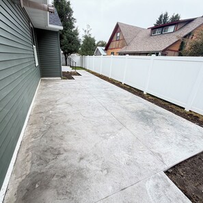 Un patio trasero residencial con un patio de hormigón, una valla blanca y una casa con revestimiento gris y techo rojo.