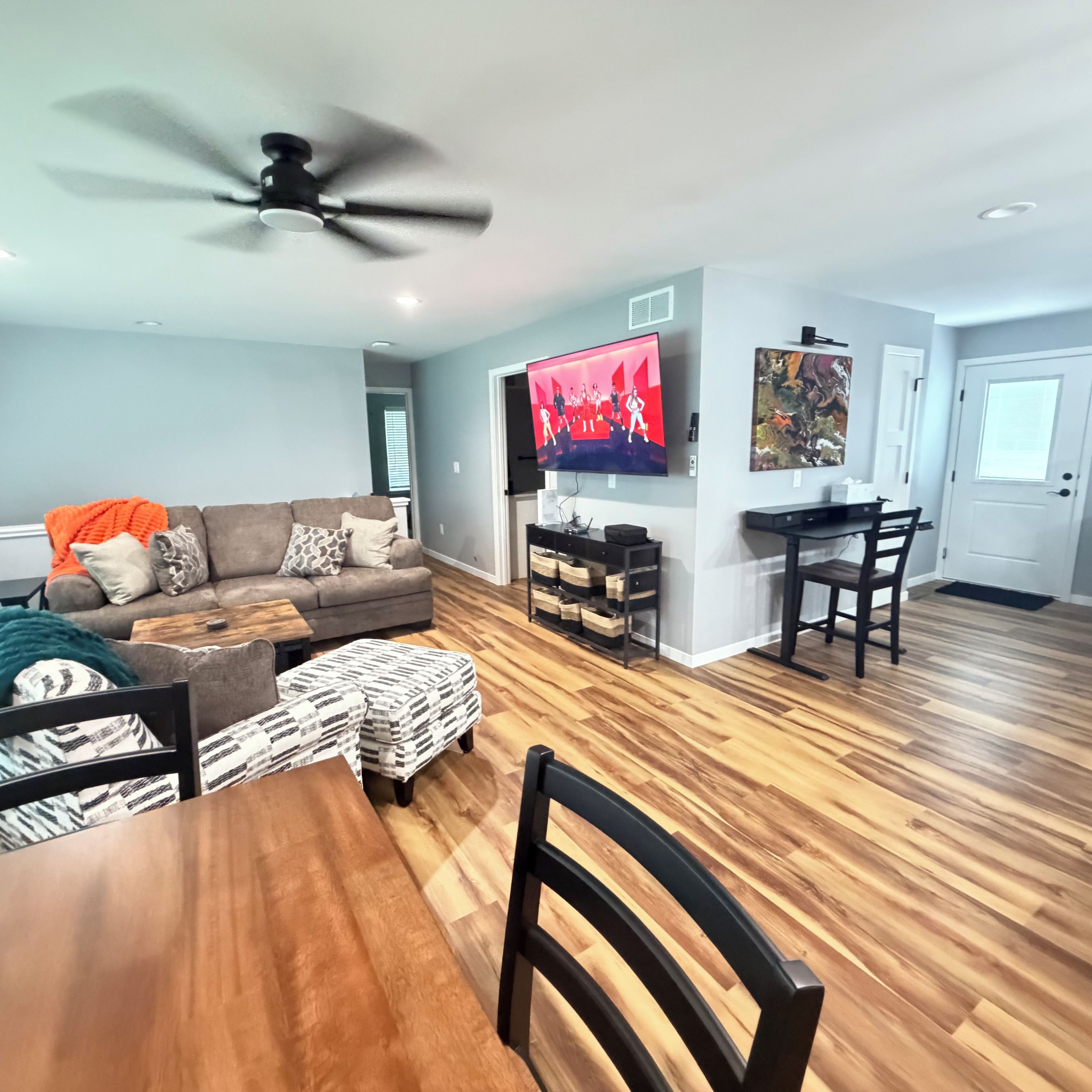 A living room with a sofa, ottoman, wooden dining table, chairs, a ceiling fan, a TV, and a painting on the wall.