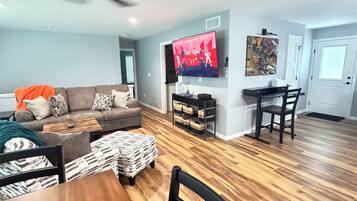 A living room with a sofa, ottoman, wooden dining table, chairs, a ceiling fan, a TV, and a painting on the wall.