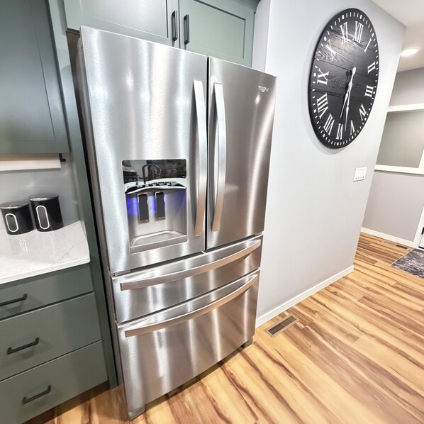A stainless steel refrigerator with a digital display, a wooden floor, and a wall clock.