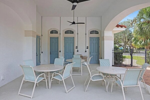 A covered patio with white walls, blue doors, and a ceiling fan. There are multiple tables and chairs arranged for outdoor dining.